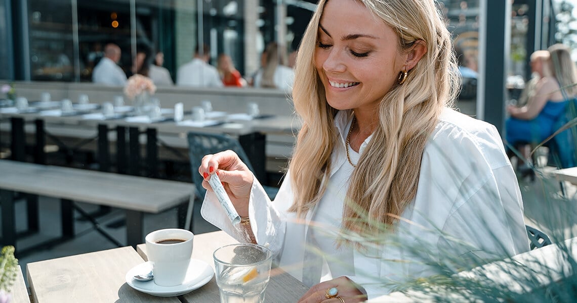 žena sedící venku u stolu v kavárně, sype si kolagen do kávy